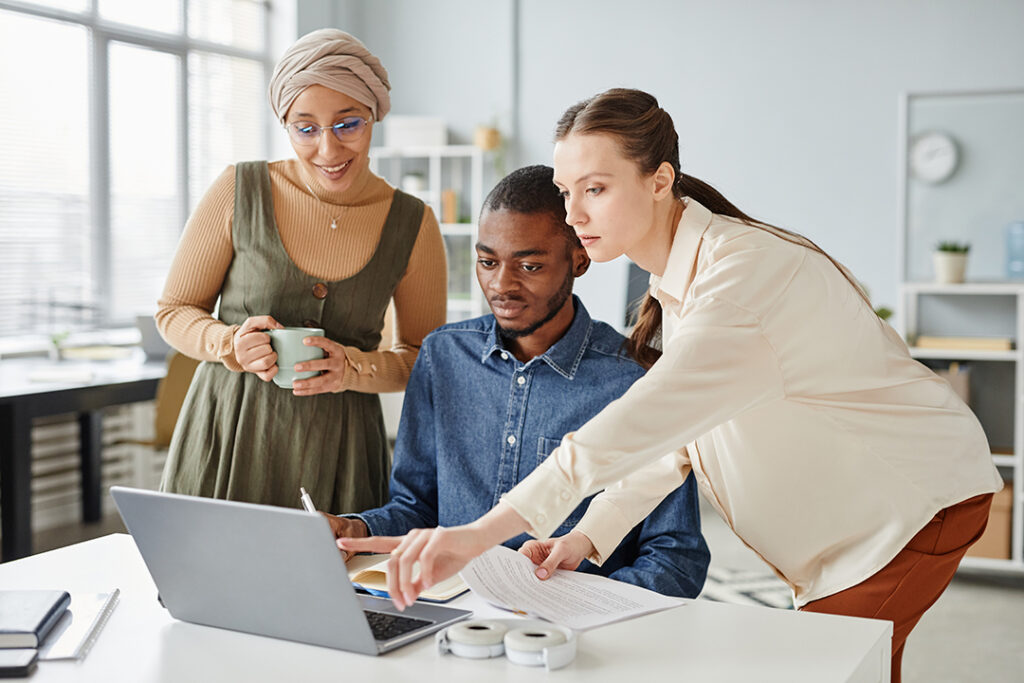 Three colleagues discuss digital marketing strategies around a laptop in a bright office, one pointing at the screen.