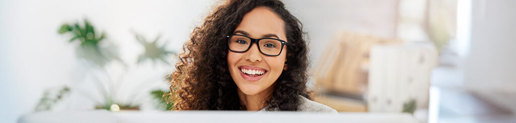 A woman with curly hair and glasses smiles while working on diy marketing at a desk.