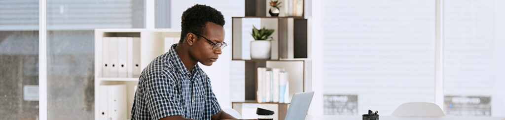 Man wearing glasses works on a laptop at a desk in a bright, modern office with shelves and plants in the background.