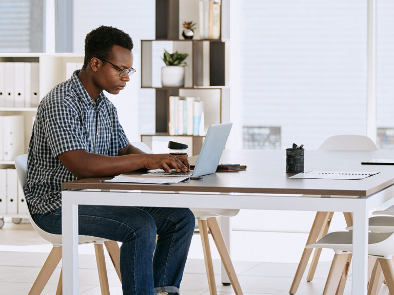 Man wearing glasses working on a laptop at a modern office desk with shelves and plants in the background.