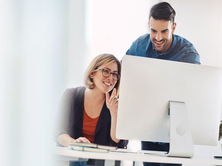 Two people look at a computer monitor together in an office setting, appearing engaged and focused on the screen.