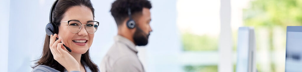 Two customer service representatives wearing headsets work at computers in a modern office setting.