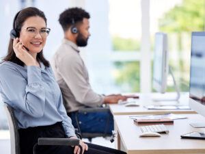 Two customer service representatives wearing headsets work at desks with computers in a bright office.