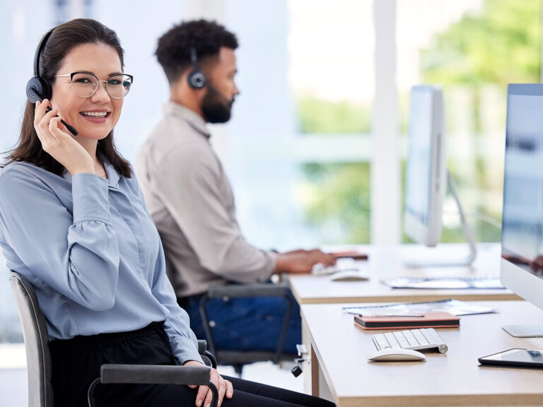 Two customer service representatives wearing headsets work at desks with computers in a bright office.