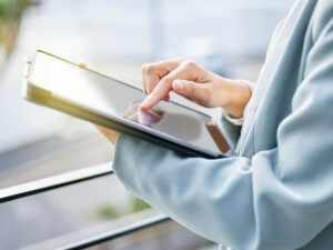 Person in a light blue blazer uses a tablet device, tapping the screen with their finger in an outdoor setting.