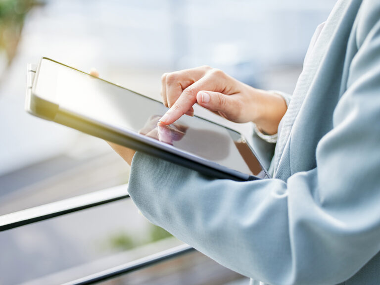 Person in a light blue blazer uses a tablet device, tapping the screen with their finger in an outdoor setting.