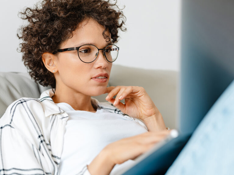 Woman with curly hair and glasses on a couch, focused on her laptop, researching lead generation mistakes.