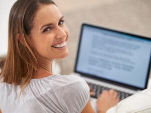 Woman smiling at the camera while typing on a laptop to improve SEO with blogging.