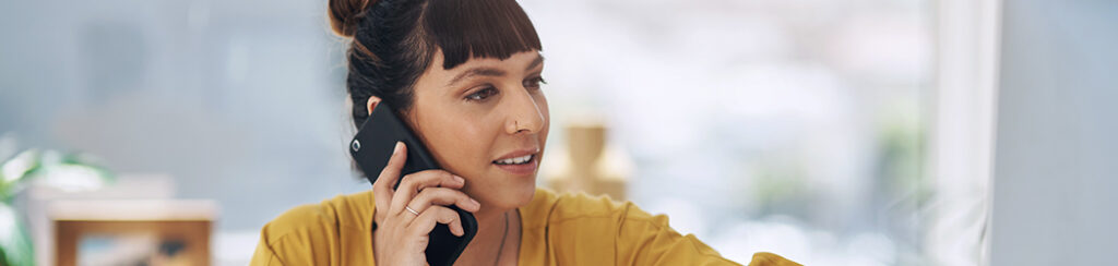 Woman with bangs wearing a yellow top talks on a smartphone while looking at something offscreen.