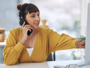 Woman in a yellow blouse talks on the phone while working at a computer in a bright office setting.