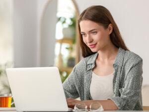 Woman with long brown hair uses a laptop at a desk indoors, with a cup and glasses in the foreground.