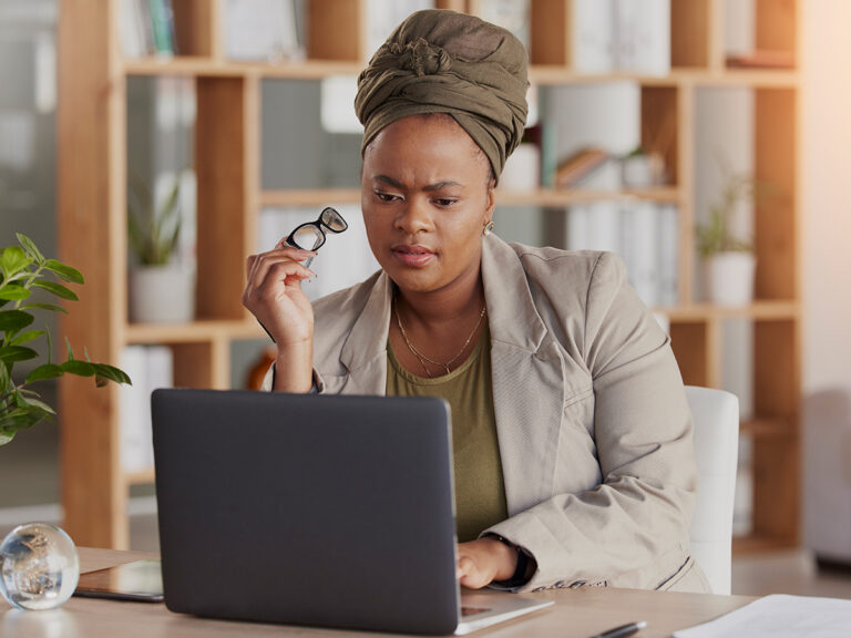 A woman in a headwrap researches "is SEO worth it" on her laptop, holding eyeglasses