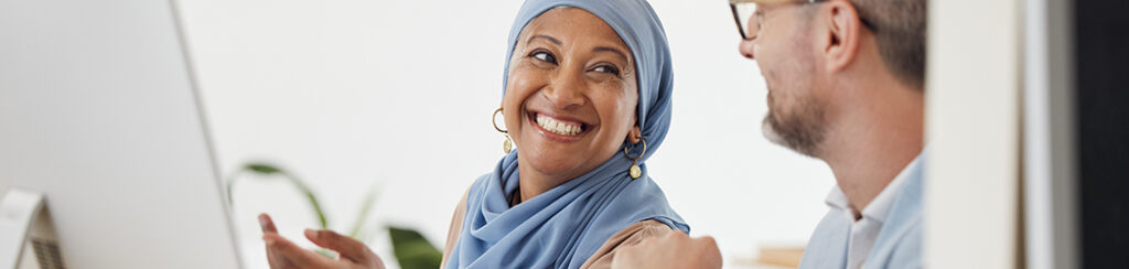 A woman in a blue headscarf smiles at a man as they discuss marketing partnerships at a computer.