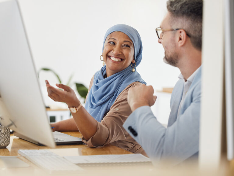 Two colleagues smile and chat about marketing partnerships while working at a desk with a computer.