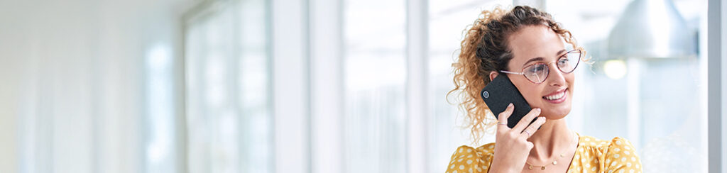 Woman with curly hair and glasses smiles while talking about high quality leads on her smartphone near a window.