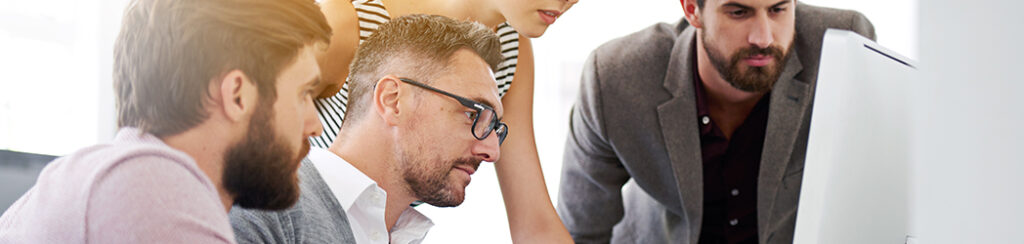 Four people gather around a monitor, focused on ensuring marketing consistency in their work-related discussion.