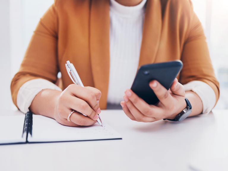 Woman writing notes while checking phone, planning lead nurture follow-up strategy for local business growth.